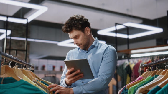 Consultor de campo focado, vestindo camisa social azul clara, segurando um tablet enquanto inspeciona peças de roupa em uma arara de madeira dentro de uma loja de varejo com iluminação moderna no teto. Em primeiro plano, à esquerda, destaca-se uma camiseta pendurada na vibrante cor verdigris. A imagem ilustra a execução prática da auditoria de franquias, mostrando a visita presencial do consultor e a automação do processo através de um checklist digital para garantir o padrão da marca.