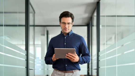 Gestor corporativo de óculos e camisa azul-escura caminhando de forma focada pelo corredor de um escritório moderno, enquanto analisa dados em um tablet. O corredor possui paredes de vidro com faixas horizontais foscas em um sutil tom verdigris. A imagem ilustra o monitoramento de indicadores, a tomada de decisão e a etapa de verificação (Check) do Ciclo PDCA na gestão de operações.