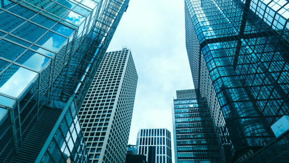 Visão em ângulo baixo (contra-plongée) apontando para o céu, mostrando imponentes arranha-céus corporativos com fachadas geométricas e espelhadas. O reflexo dos vidros dos edifícios domina a fotografia com um tom marcante e imponente de verdigris. A imagem ilustra o crescimento empresarial, a escala de negócios e o sucesso na expansão, conectando-se ao guia sobre como abrir uma filial, entender os modelos de expansão e estruturar a arquitetura operacional da nova unidade.