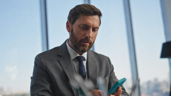 Gestor executivo de barba, vestindo terno cinza e gravata azul, focado enquanto acessa informações em um smartphone com capinha na cor verdigris em um escritório moderno com grandes janelas de vidro. Em primeiro plano, elementos de escritório aparecem desfocados. A imagem ilustra a eficácia dos canais mobile na comunicação interna para redes, garantindo que a informação estratégica chegue de forma rápida e sem ruídos aos colaboradores e equipes distribuídas.
