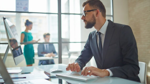 Homem de terno escuro, barba e óculos, trabalhando focado no teclado de um computador de mesa em um escritório bem iluminado. Através da divisória de vidro ao fundo, observa-se a movimentação do ambiente corporativo, com destaque para uma profissional em pé vestindo um vestido na cor verdigris. A imagem ilustra o trabalho analítico de retaguarda, a revisão remota de dados e o planejamento estratégico necessários para estruturar um programa moderno de consultoria de campo em franquias, transformando o auditor em um verdadeiro coach de negócios.