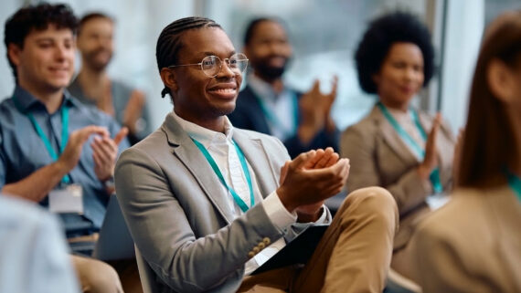 Plateia diversificada de profissionais sorridentes sentados em um auditório corporativo, aplaudindo entusiasticamente. Em primeiro plano, um executivo negro de óculos e blazer cinza lidera os aplausos. Todos os participantes visíveis usam cordões de crachá corporativos padronizados na cor verdigris. A imagem ilustra o alto nível de engajamento, a retenção de talentos e o alinhamento estratégico alcançados em uma convenção de franquia de sucesso, evento fundamental para sincronizar a cultura da rede.