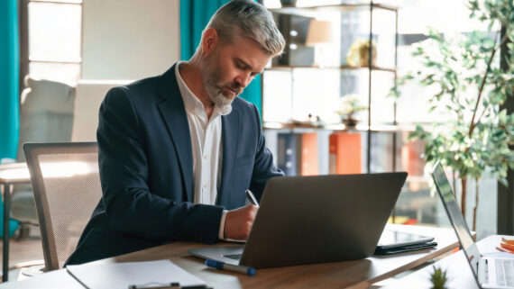 Gestor sênior de terno escuro sentado à mesa de um escritório moderno, escrevendo anotações à mão de forma focada ao lado de seu notebook aberto. Ao fundo, longas cortinas em um vibrante tom verdigris emolduram a janela do ambiente, que também conta com plantas e estantes. A imagem ilustra a etapa de planejamento, criação e documentação de um Procedimento Operacional Padrão (POP), estruturando o passo a passo seguro para a execução de atividades rotineiras.