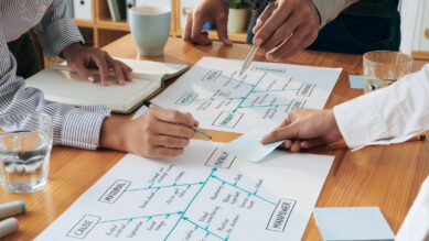 Mãos de uma equipe de profissionais reunidos ao redor de uma mesa de madeira, utilizando canetas para analisar dois Diagramas de Ishikawa (Espinha de Peixe) desenhados no papel. As setas estruturais de causa e efeito dos diagramas estão traçadas na cor verdigris, organizando as categorias de análise. A cena ilustra a aplicação prática da ferramenta em equipe para identificar a causa raiz de um problema utilizando o método dos 6Ms.
