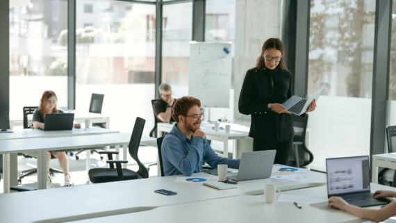 Ambiente de escritório open space claro e moderno com profissionais trabalhando. Em primeiro plano, uma mulher em pé de blusa preta compartilha informações na tela do seu notebook com um colega ruivo de óculos, que sorri engajado enquanto olha para a tela. Sobre a mesa branca, documentos impressos exibem um gráfico de pizza com uma fatia destacada na cor verdigris. Ao fundo, outros colaboradores focados em seus computadores. A imagem ilustra o alinhamento, a colaboração e as estratégias de endomarketing no trabalho híbrido, essenciais para engajar equipes, tratar o colaborador como cliente interno e reter talentos.