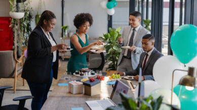 Quatro profissionais sorridentes celebrando juntos em um escritório moderno ao redor de uma mesa de reunião com bolo de chocolate, frutas, caixas de presente e um notebook. O ambiente festivo é decorado com balões, destacando-se fortemente os da cor verdigris, mesmo tom elegante do vestido usado por uma das colaboradoras no centro da cena. A imagem ilustra uma ação prática de endomarketing, demonstrando a valorização do colaborador como cliente interno, a melhoria do clima organizacional e o uso de estratégias para engajar e reter talentos.