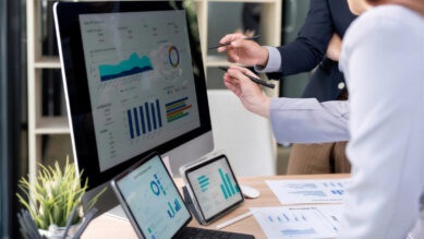 Dois profissionais em um escritório apontando com canetas para um painel de dados em um monitor de computador. A tela exibe gráficos de barras, pizza e um gráfico de área na cor verdigris. Sobre a mesa de madeira, há tablets, relatórios impressos e uma planta, ilustrando a análise de dados e a aplicação prática das ferramentas da qualidade na gestão de processos.