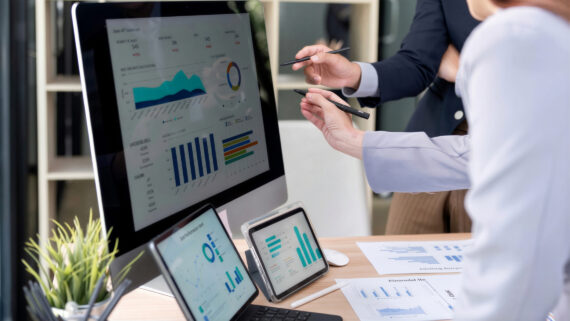 Dois profissionais em um escritório apontando com canetas para um painel de dados em um monitor de computador. A tela exibe gráficos de barras, pizza e um gráfico de área na cor verdigris. Sobre a mesa de madeira, há tablets, relatórios impressos e uma planta, ilustrando a análise de dados e a aplicação prática das ferramentas da qualidade na gestão de processos.