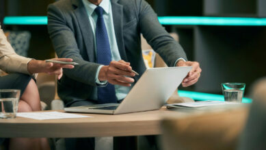 Dois profissionais em uma reunião corporativa analisando dados na tela de um notebook sobre uma mesa de madeira. O homem de terno cinza e gravata azul segura o computador e uma caneta, enquanto a colega, vestindo um blazer claro, aponta para a tela com outra caneta. Sobre a mesa, há relatórios impressos e copos com água. Ao fundo, uma moderna iluminação em fita de LED na cor verdigris destaca o ambiente. A imagem ilustra o trabalho em equipe para a avaliação de riscos, análise de severidade e planejamento de ações preventivas utilizando a metodologia FMEA.