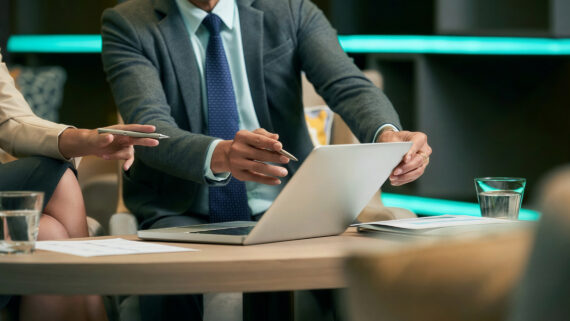 Dois profissionais em uma reunião corporativa analisando dados na tela de um notebook sobre uma mesa de madeira. O homem de terno cinza e gravata azul segura o computador e uma caneta, enquanto a colega, vestindo um blazer claro, aponta para a tela com outra caneta. Sobre a mesa, há relatórios impressos e copos com água. Ao fundo, uma moderna iluminação em fita de LED na cor verdigris destaca o ambiente. A imagem ilustra o trabalho em equipe para a avaliação de riscos, análise de severidade e planejamento de ações preventivas utilizando a metodologia FMEA.