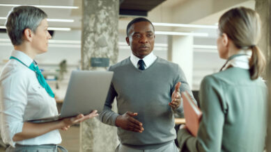 Três profissionais conversando em pé em um escritório corporativo moderno. À esquerda, uma mulher segura um notebook e veste uma camisa branca com um lenço verdigris no pescoço. No centro, um gestor de suéter cinza gesticula durante uma explicação, enquanto outra mulher à direita escuta segurando um caderno, ilustrando a comunicação, o alinhamento de equipe e as práticas de gestão administrativa.