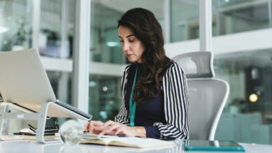 Gestora trabalhando concentrada em um teclado conectado a um notebook sobre suporte ergonômico, em um escritório corporativo envidraçado. Ela usa uma camisa listrada com um cordão de crachá na cor verdigris, mesma cor da capa do tablet apoiado sobre a mesa ao lado de um caderno. A cena ilustra o foco no monitoramento de fluxos de trabalho, produtividade e a aplicação prática da gestão de processos (BPM).