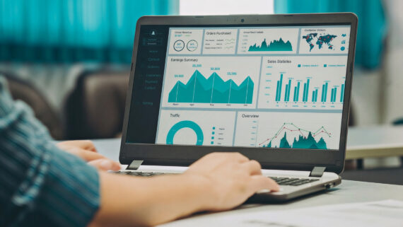 Visão sobre o ombro de um profissional digitando em um notebook sobre uma mesa clara. A tela exibe um painel de controle (dashboard) gerencial detalhado com gráficos de área, barras, indicadores circulares e um mapa-múndi, todos destacados na cor verdigris, harmonizando com as cortinas ao fundo do ambiente. A imagem ilustra o monitoramento em tempo real dos 15 indicadores essenciais (KPIs), a análise de performance financeira e comercial, e a governança baseada em dados para redes de franquias.