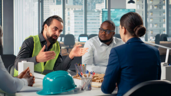 Quatro profissionais reunidos em uma mesa de escritório com vista para a cidade. Um homem de barba vestindo colete refletivo amarelo gesticula enquanto explica um projeto para a equipe. Sobre a mesa, há plantas arquitetônicas, porta-canetas, uma maquete e um capacete de segurança na cor verdigris em primeiro plano. A imagem ilustra o alinhamento de equipes e o planejamento corporativo para garantir a conformidade com as Normas Regulamentadoras (NRs).