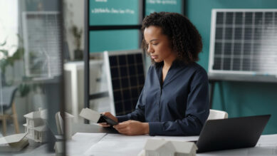 Engenheira de camisa azul-escura sentada à mesa do escritório, analisando anotações com atenção. Sobre a mesa há plantas de projeto, maquetes de casas brancas e um notebook aberto. Ao fundo, painéis solares estão apoiados perto de uma parede na cor verdigris. A imagem ilustra o planejamento técnico, a análise de escopo e a emissão de uma Ordem de Serviço (OS) para a execução padronizada de trabalhos.