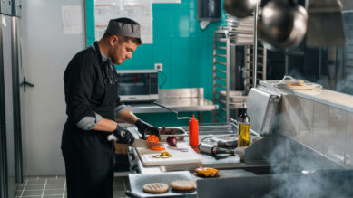 Chef vestindo uniforme e chapéu pretos, além de luvas escuras, fatiando um tomate sobre uma tábua de corte branca em uma cozinha industrial de aço inox. Ao fundo, a parede do restaurante é revestida com azulejos na cor verdigris. Em primeiro plano, fumaça sobe de uma chapa quente onde hambúrgueres são preparados. A imagem ilustra a execução rigorosa de padrões de qualidade e a padronização de processos no setor de food service, essencial para manter a consistência em redes e franquias.