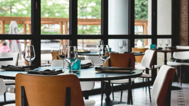 Interior de um restaurante moderno e bem iluminado por amplas janelas de vidro. Em primeiro plano, mesas redondas escuras estão meticulosamente arrumadas com taças de vinho, guardanapos de tecido escuro e copos decorativos na cor verdigris. As cadeiras mesclam acabamentos em madeira e estofados brancos. A imagem ilustra a aplicação prática da padronização visual de unidades, demonstrando a consistência do layout, da ambientação e da identidade visual no ponto de venda para fortalecer a marca e evitar o brand drift.