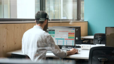 Visão através de uma divisória de vidro de um profissional focado, trabalhando em um computador em um escritório moderno com vista para a rua. Ele analisa um dashboard repleto de gráficos e indicadores na tela, representando o módulo de NPS. O ambiente conta com mesas brancas, cadeiras pretas e uma parede de destaque na cor verdigris à direita. A cena ilustra o monitoramento de métricas, o cálculo e a interpretação de resultados de uma pesquisa de satisfação.