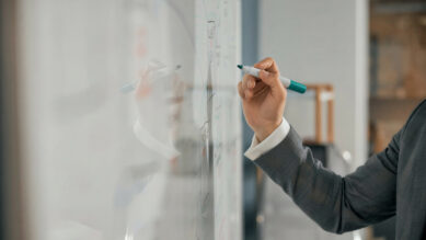 Braço de um profissional vestindo terno cinza escrevendo em um quadro branco com um marcador na cor verdigris. O quadro possui diversas anotações e esquemas visuais, ilustrando a etapa de planejamento e a aplicação de metodologias para estruturar um plano de ação.