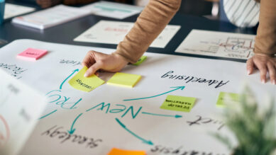 Pessoa debruçada sobre uma mesa de escritório colando um post-it amarelo em um grande papel branco. O papel mostra o esboço visual de um planejamento estratégico com várias anotações e setas direcionais desenhadas na cor verdigris, ilustrando o passo a passo prático de como fazer um plano de trabalho.