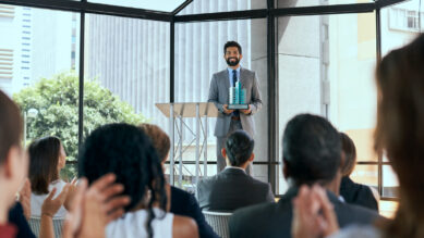Visão a partir da plateia de uma cerimônia corporativa, mostrando pessoas desfocadas em primeiro plano aplaudindo. No palco ao centro, um executivo sorridente de terno cinza e gravata azul está posicionado ao lado de um púlpito, segurando com orgulho um troféu moderno que destaca colunas verticais na cor verdigris. Ao fundo, um grande painel de vidro revela a arquitetura urbana da cidade iluminada pelo sol. A cena ilustra o momento do reconhecimento simbólico em uma cerimônia anual de premiação de franqueados, celebrando o engajamento e a aderência das unidades ao padrão da marca.