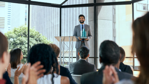 Visão a partir da plateia de uma cerimônia corporativa, mostrando pessoas desfocadas em primeiro plano aplaudindo. No palco ao centro, um executivo sorridente de terno cinza e gravata azul está posicionado ao lado de um púlpito, segurando com orgulho um troféu moderno que destaca colunas verticais na cor verdigris. Ao fundo, um grande painel de vidro revela a arquitetura urbana da cidade iluminada pelo sol. A cena ilustra o momento do reconhecimento simbólico em uma cerimônia anual de premiação de franqueados, celebrando o engajamento e a aderência das unidades ao padrão da marca.