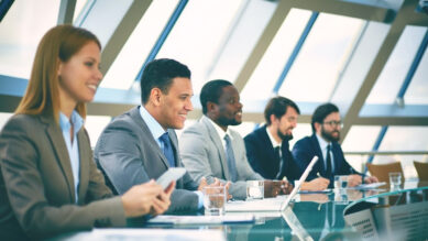 Grupo de cinco executivos diversificados e sorridentes sentados lado a lado em uma mesa de conferência de vidro em um escritório moderno. Eles olham com atenção e aprovação para a direita, acompanhando uma apresentação. O ambiente possui grandes janelas com vigas diagonais e a cena é banhada por uma iluminação que reflete um tom marcante de verdigris, especialmente na superfície da mesa. A imagem ilustra o alinhamento da liderança e a aprovação de métricas claras para criar e gerir o ranking de franqueados, reduzindo a variância e avaliando a performance da rede.