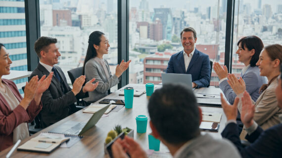 Grupo diverso de executivos em uma sala de reunião corporativa moderna, com amplas janelas de vidro e vista panorâmica para a cidade. Eles estão sentados ao redor de uma mesa de conferência, sorrindo e aplaudindo um colega ao centro. Sobre a mesa, ao lado de notebooks e documentos, destacam-se vários copos térmicos na cor verdigris. A imagem de comemoração e aplausos ilustra o reconhecimento de alta performance, a premiação por excelência e o engajamento gerado por um ranking de franqueados bem estruturado para gerir o desempenho da rede.