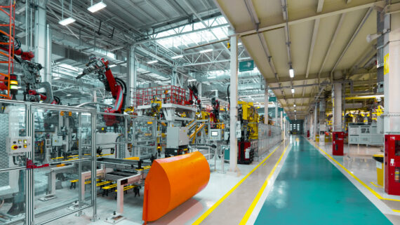 Amplo corredor de uma fábrica automotiva moderna e automatizada. À esquerda, braços robóticos vermelhos operam isolados por grades de segurança transparentes. O piso do corredor de circulação principal, delimitado por faixas de segurança amarelas, é pintado em um vibrante tom verdigris. A imagem ilustra o controle de perigos físicos e mecânicos em um ambiente industrial, demonstrando na prática a prevenção de acidentes e a aplicação do Gerenciamento de Riscos Ocupacionais (GRO).