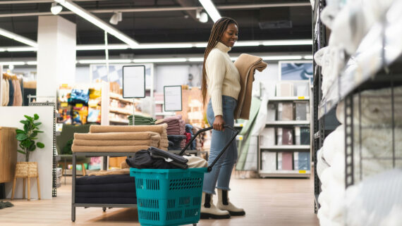 Uma mulher sorridente com tranças longas, vestindo um suéter creme e jeans, fazendo compras em uma loja de artigos para o lar bem iluminada. Ela está segurando um cobertor de lã enrolado e puxando um carrinho de compras com rodas na cor verdigris. Há também um pequeno vaso de planta com folhagem verde ao lado de uma pilha de cobertores. Prateleiras cheias de produtos de cama e banho brancos e em tons neutros, junto com cartazes de preços brancos, alinham os corredores, ilustrando a execução de estratégias de trade marketing no ponto de venda (PDV).
