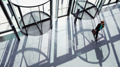 Visão superior (plongeé) de uma profissional segurando uma pasta executiva, vestindo um terninho escuro e uma blusa na cor verdigris, passando por uma grande porta giratória de vidro na entrada de um edifício corporativo moderno. A luz do sol projeta longas sombras no piso claro. A imagem utiliza a metáfora visual da porta giratória para ilustrar a evasão de talentos, conectando-se ao guia sobre como aplicar onboarding estruturado e engajamento para reduzir o turnover e a rotatividade de funcionários em redes de franquias.