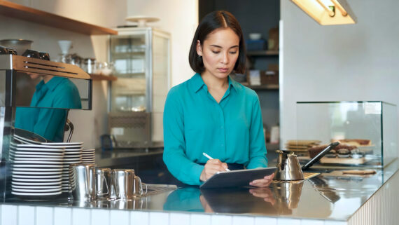 Gestora focada, vestindo uma camisa de botão na cor verdigris, utilizando uma caneta digital e um tablet para realizar verificações atrás do balcão de aço inox de uma cafeteria moderna. Ao seu lado, há uma máquina de café expresso, pilhas de xícaras brancas e, ao fundo, prateleiras e uma vitrine. A cena ilustra a condução de uma vistoria técnica presencial, como a sanitária ou de qualidade, documentando as condições do estabelecimento para garantir a conformidade operacional.