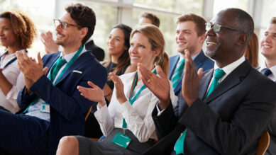 Público diversificado de profissionais corporativos sentados em um auditório moderno e bem iluminado por janelas de vidro. Eles sorriem e aplaudem entusiasticamente em direção ao palco. Destaca-se a forte padronização visual dos participantes: os homens vestem ternos com gravatas na cor verdigris, enquanto a mulher ao centro, de camisa branca, utiliza um cordão e crachá de identificação no mesmo tom verdigris. A imagem ilustra a energia, o engajamento e o alinhamento estratégico gerados por uma convenção de franquia bem executada, evento essencial para sincronizar a cultura da rede.