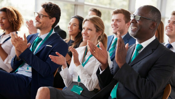 Público diversificado de profissionais corporativos sentados em um auditório moderno e bem iluminado por janelas de vidro. Eles sorriem e aplaudem entusiasticamente em direção ao palco. Destaca-se a forte padronização visual dos participantes: os homens vestem ternos com gravatas na cor verdigris, enquanto a mulher ao centro, de camisa branca, utiliza um cordão e crachá de identificação no mesmo tom verdigris. A imagem ilustra a energia, o engajamento e o alinhamento estratégico gerados por uma convenção de franquia bem executada, evento essencial para sincronizar a cultura da rede.