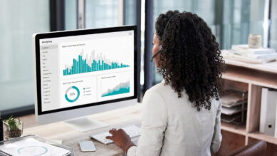 Visão sobre o ombro de uma profissional negra com cabelos cacheados, usando headset e blusa clara, sentada trabalhando em frente a um grande monitor em um escritório moderno. A tela do computador exibe um painel analítico com destaque para um grande histograma cujas barras contíguas brilham na cor verdigris, além de um gráfico de rosca no mesmo tom. A imagem ilustra a análise visual e a interpretação da distribuição de dados, conectando-se ao guia prático sobre como usar essa ferramenta da qualidade para diagnosticar a estabilidade de processos e métricas operacionais.
