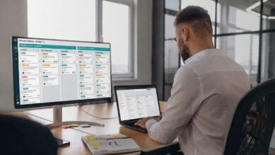 Visão sobre o ombro de um profissional de camisa branca trabalhando em sua mesa com um notebook e um monitor ultrawide. A tela do grande monitor exibe um quadro Kanban digital organizando tarefas em formato de cartões distribuídos em colunas. O cabeçalho superior do software e as tags das colunas destacam-se na vibrante cor verdigris. A imagem ilustra de forma prática o funcionamento do sistema visual de gestão de fluxo de trabalho, conectando-se ao guia prático sobre como aplicar o Kanban e organizar processos corporativos.