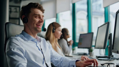 Profissional de suporte corporativo sorridente, vestindo camisa social azul clara e headset, digitando no teclado enquanto olha para a tela do computador. Ao fundo, em um ambiente de escritório bem iluminado, outras colaboradoras também usam headsets em suas estações de trabalho. As amplas janelas do local possuem esquadrias metálicas destacadas na cor verdigris. A imagem ilustra a equipe da franqueadora interagindo com a rede, momento chave onde é captada a percepção de atendimento para alimentar o NES (Net Emotional Score) e mapear a satisfação e o sentimento de cada unidade.