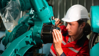 Visão aproximada de um jovem técnico industrial vestindo capacete branco e uniforme vermelho, focado enquanto realiza a manutenção manual nas engrenagens de um grande braço robótico. A robusta estrutura do maquinário domina a imagem, destacando-se intensamente na vibrante cor verdigris. A fotografia ilustra a prática no chão de fábrica da Manutenção Produtiva Total (TPM), conectando-se ao método de envolver os colaboradores na operação para garantir zero quebra, evitar defeitos e elevar o indicador OEE.