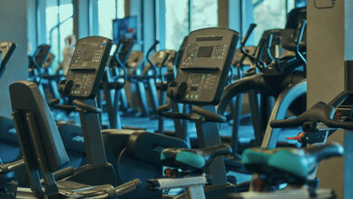 Visão interna de uma academia moderna, sofisticada e vazia, exibindo uma fileira perfeitamente alinhada de equipamentos de cardio de última geração. Em primeiro plano, destacam-se bicicletas ergométricas cujos detalhes de acabamento nos assentos brilham intensamente na cor verdigris. Ao fundo, veem-se máquinas elípticas sob uma iluminação ambiente elegante e profissional. A fotografia ilustra uma infraestrutura de alto nível pronta para operação, conectando-se ao guia completo sobre gestão de academia, focado em utilizar dados e KPIs para profissionalizar o controle financeiro, otimizar a equipe e aumentar a retenção de alunos.