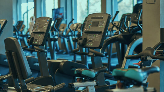 Visão interna de uma academia moderna, sofisticada e vazia, exibindo uma fileira perfeitamente alinhada de equipamentos de cardio de última geração. Em primeiro plano, destacam-se bicicletas ergométricas cujos detalhes de acabamento nos assentos brilham intensamente na cor verdigris. Ao fundo, veem-se máquinas elípticas sob uma iluminação ambiente elegante e profissional. A fotografia ilustra uma infraestrutura de alto nível pronta para operação, conectando-se ao guia completo sobre gestão de academia, focado em utilizar dados e KPIs para profissionalizar o controle financeiro, otimizar a equipe e aumentar a retenção de alunos.