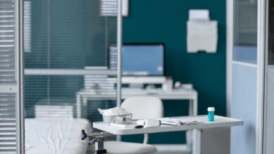 Visão interna de um consultório médico moderno e organizado. Em primeiro plano, uma mesa auxiliar contém instrumentos médicos, um termômetro e um pequeno frasco; à esquerda, parte de uma maca de exames com lençol branco. Ao fundo, separada por divisórias de vidro com persianas, vê-se uma estação de trabalho administrativa com computador e cadeira. A parede principal do consultório e o frasco sobre a mesa destacam-se fortemente na cor verdigris. A fotografia ilustra a infraestrutura de saúde em operação, conectando-se ao guia estratégico sobre gestão de clínica para estruturar pilares operacionais, garantir o controle financeiro, monitorar KPIs e manter o compliance regulatório da Anvisa e CFM.