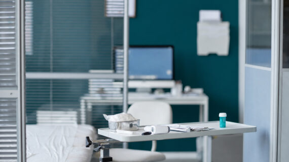 Visão interna de um consultório médico moderno e organizado. Em primeiro plano, uma mesa auxiliar contém instrumentos médicos, um termômetro e um pequeno frasco; à esquerda, parte de uma maca de exames com lençol branco. Ao fundo, separada por divisórias de vidro com persianas, vê-se uma estação de trabalho administrativa com computador e cadeira. A parede principal do consultório e o frasco sobre a mesa destacam-se fortemente na cor verdigris. A fotografia ilustra a infraestrutura de saúde em operação, conectando-se ao guia estratégico sobre gestão de clínica para estruturar pilares operacionais, garantir o controle financeiro, monitorar KPIs e manter o compliance regulatório da Anvisa e CFM.
