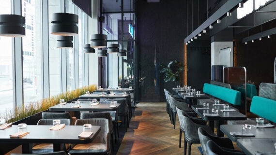 Visão interna do salão de um restaurante moderno, sofisticado e vazio, com as mesas perfeitamente arrumadas com copos e guardanapos para o início da operação. O ambiente possui amplas janelas de vidro, piso de madeira e luminárias elegantes. À direita, os estofados dos assentos destacam-se intensamente na cor verdigris. A fotografia ilustra o controle de salão e o alto padrão de serviço, conectando-se ao guia estratégico sobre gestão de restaurante para profissionalizar processos financeiros, controlar o CMV, monitorar KPIs de lucratividade e aplicar checklists operacionais diários.