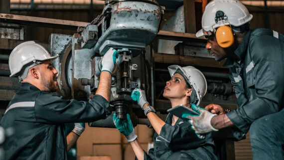 Visão em plano médio de uma equipe diversificada de manutenção industrial composta por três profissionais (uma mulher e dois homens). Eles vestem capacetes brancos e uniformes de trabalho cinza escuro, concentrados no reparo de um grande maquinário industrial. A profissional no centro utiliza um tablet para consultar dados, enquanto os colegas ajustam o equipamento mecânico. As luvas de proteção (EPI) utilizadas pela equipe destacam-se intensamente na cor verdigris. A fotografia ilustra o esforço prático para otimizar o tempo de reparo e garantir a confiabilidade dos equipamentos, conectando-se ao guia completo sobre como calcular o MTBF e o MTTR para elevar a disponibilidade dos ativos.