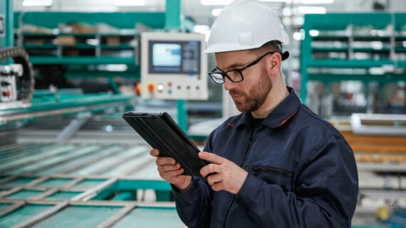 Visão em plano médio de um supervisor industrial focado, vestindo capacete branco de segurança, óculos e uniforme azul escuro, analisando dados em um tablet em suas mãos. Ao fundo, o ambiente de chão de fábrica exibe um painel de controle e grandes estruturas metálicas de maquinário industrial pintadas intensamente na cor verdigris. A fotografia ilustra a digitalização e o monitoramento em tempo real do chão de fábrica, conectando-se ao processo do Planejamento e Controle da Produção (PCP) para coordenar recursos, definir quantidades e integrar a manufatura à logística.