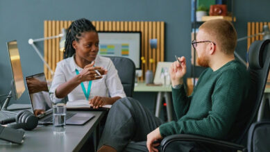 Visão em plano médio de dois profissionais engajados em uma reunião de mentoria e feedback em um escritório moderno. À esquerda, uma mulher negra com cabelos trançados, vestindo camisa branca e um cordão de crachá na cor verdigris, gesticula de forma expressiva enquanto fala. À direita, um homem branco com barba e óculos, usando um suéter em tom de verdigris escuro, escuta atentamente enquanto segura uma caneta. Sobre a mesa, veem-se notebooks, fones de ouvido e um copo de água. A imagem ilustra uma conversa focada em evolução profissional, conectando-se ao guia prático sobre como fazer e medir o Plano de Desenvolvimento Individual (PDI) para reter talentos e gerar resultados.
